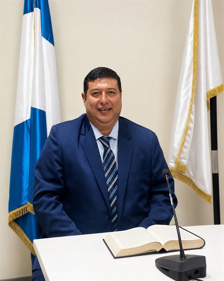 Retrato de Mario René Natareno Soto, abogado y notario, sentado ante un libro abierto entre la bandera de Guatemala y una bandera institucional.