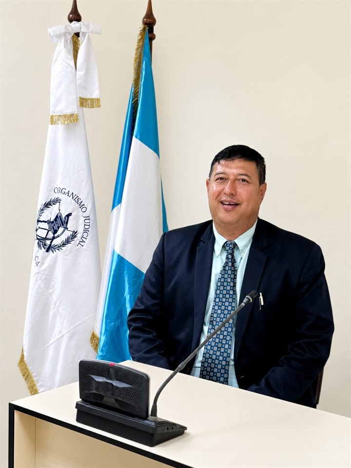 Mario René Natareno Soto, abogado y notario, sentado ante un micrófono en una sala institucional con la bandera de Guatemala y la del Organismo Judicial.